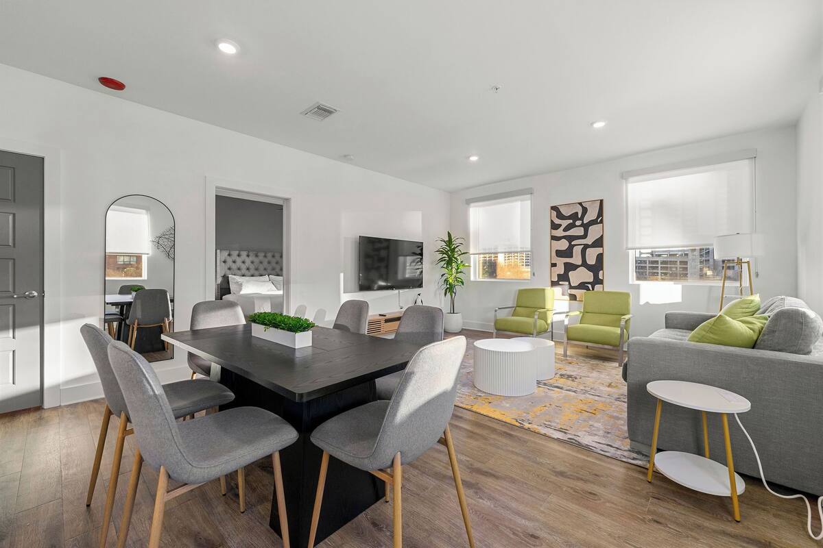 A modern living room with a grey sofa, a dining table with chairs, a TV, and a large window.