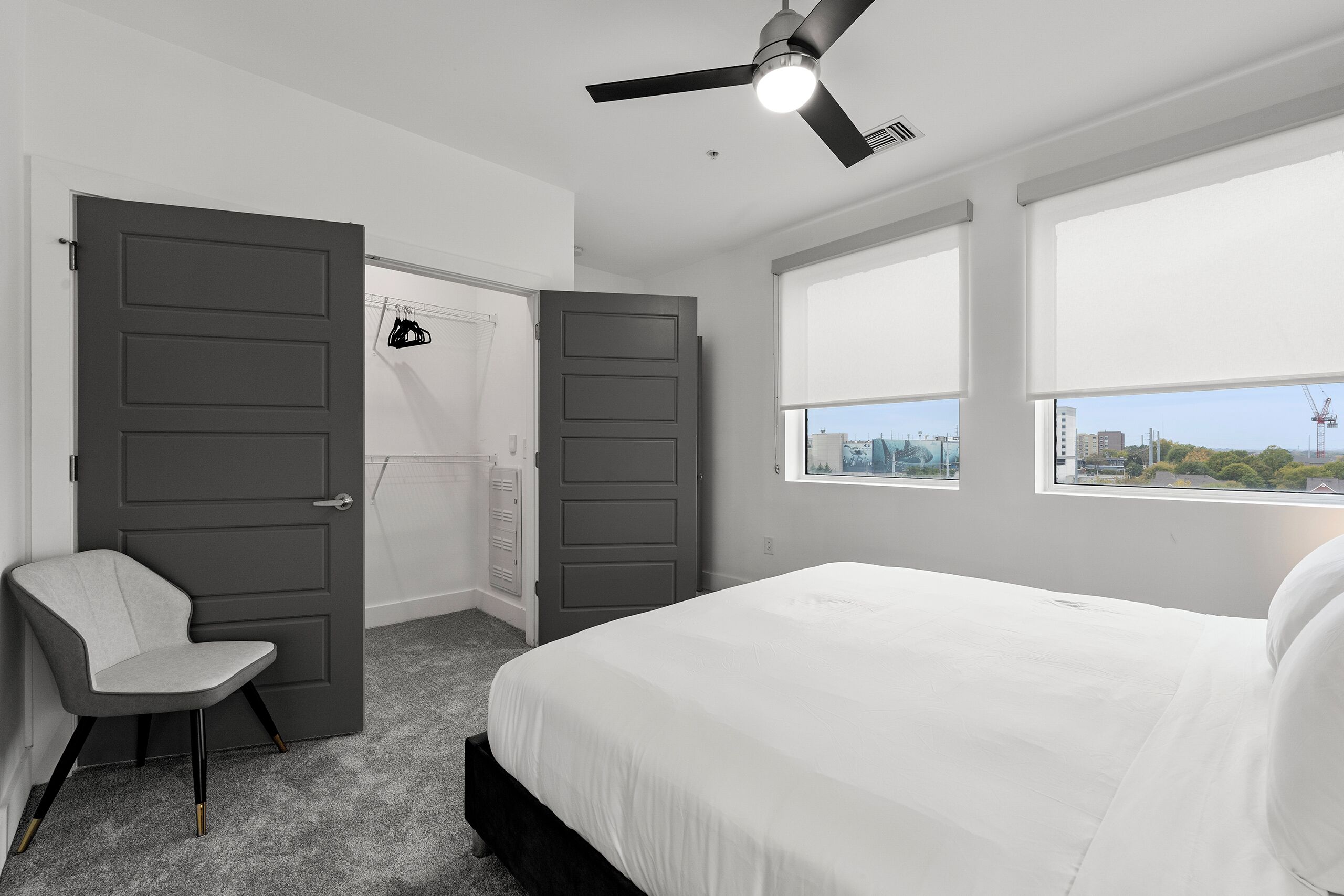 A bedroom with a bed, a chair, two doors, a ceiling fan, and a window with a city view.