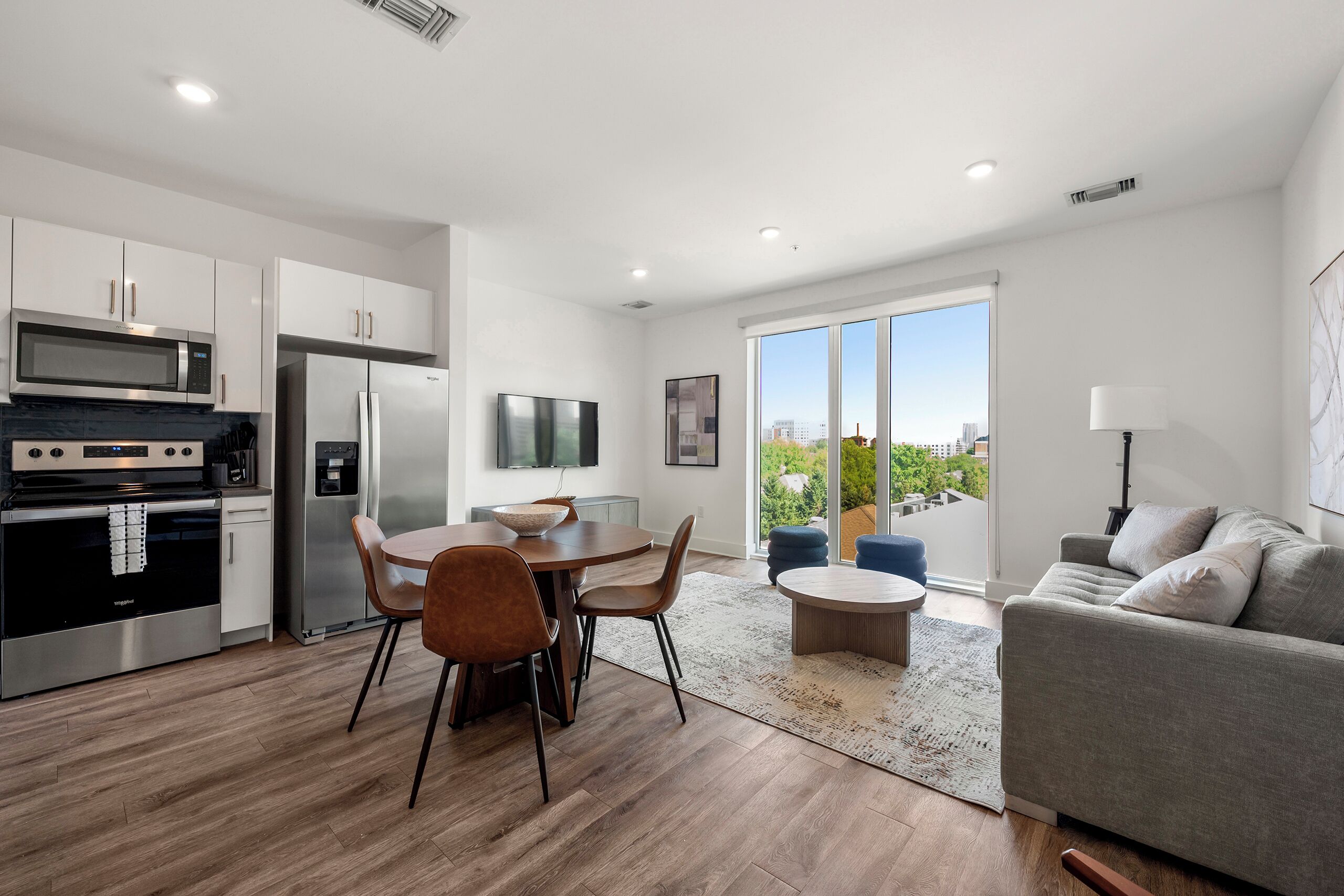 A modern kitchen with stainless steel appliances, a dining area with a round table and chairs, and a living area with a sofa and a view of the city.