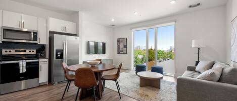 A modern kitchen with stainless steel appliances, a dining area with a round table and chairs, and a living area with a sofa and a view of the city.