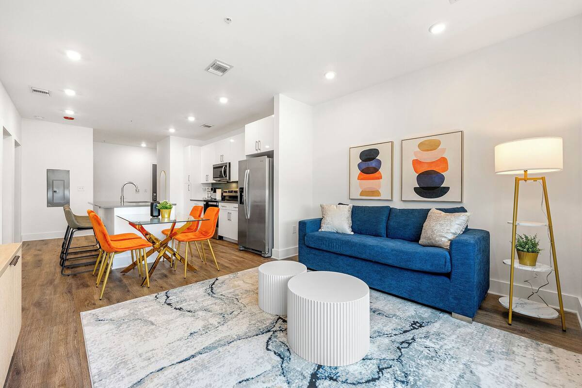 A modern living room with a blue sofa, a dining area with orange chairs, and a kitchen in the background.