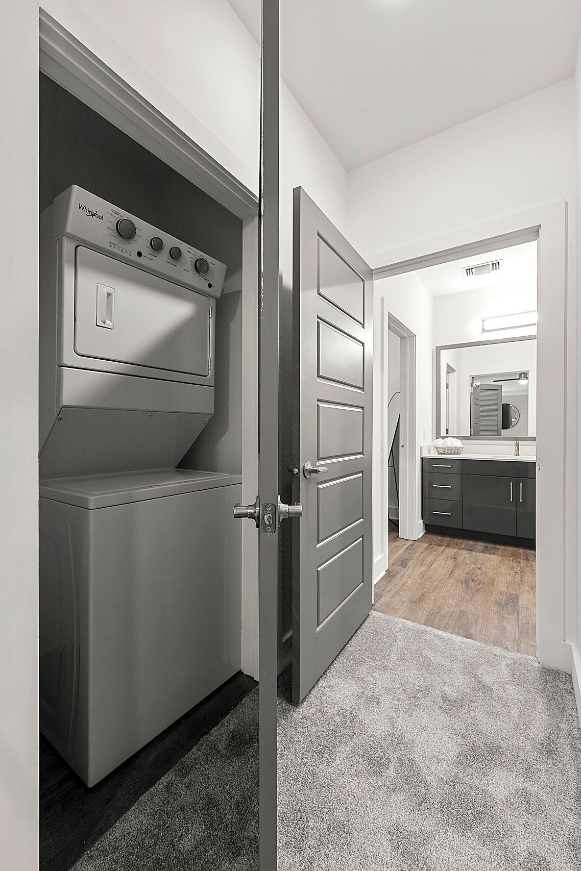 A laundry room with a front-loading washer and dryer, a gray door, and a hallway leading to a kitchen area.