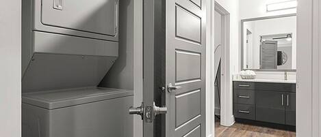 A laundry room with a front-loading washer and dryer, a gray door, and a hallway leading to a kitchen area.
