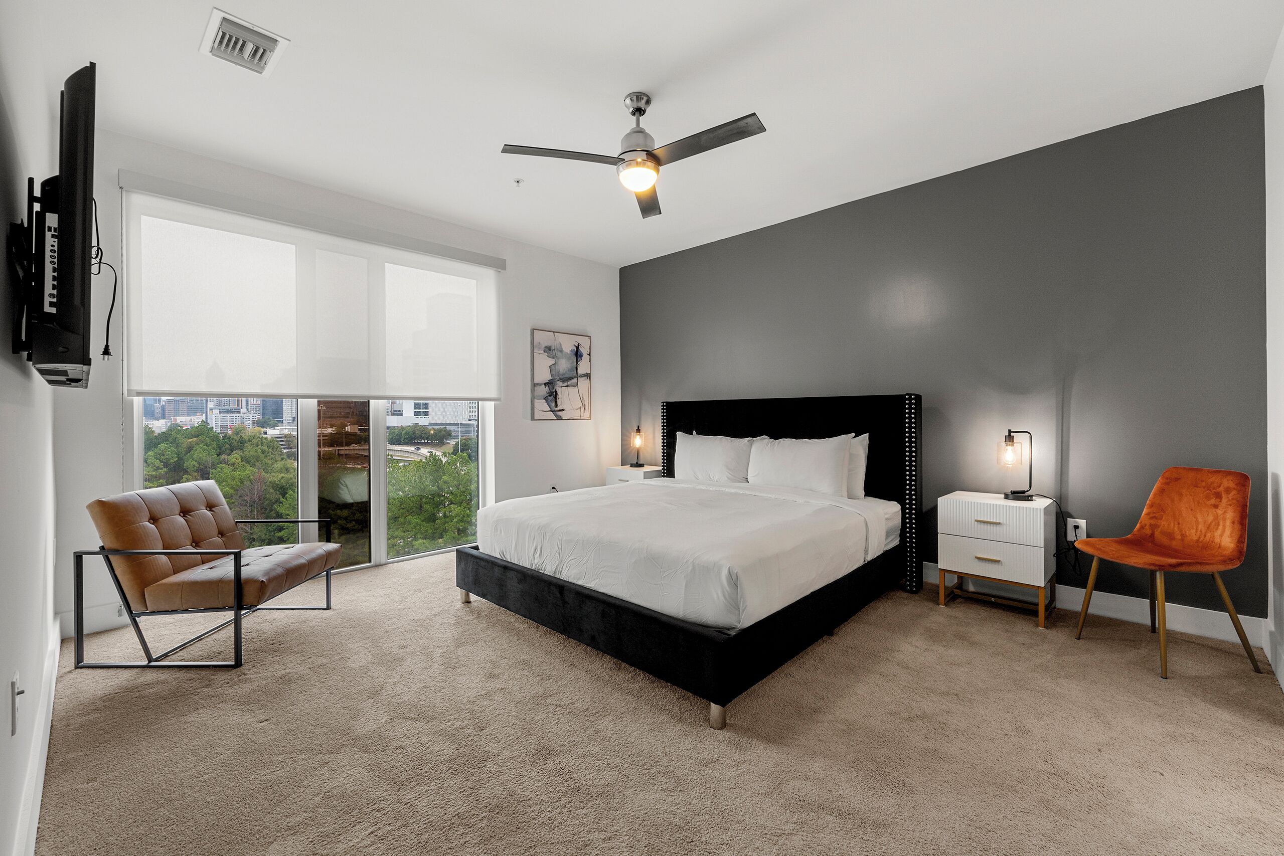 Quarto moderno com cama grande, ventilador de teto, vista para a cidade, uma cadeira e mesa de cabeceira com abajur.