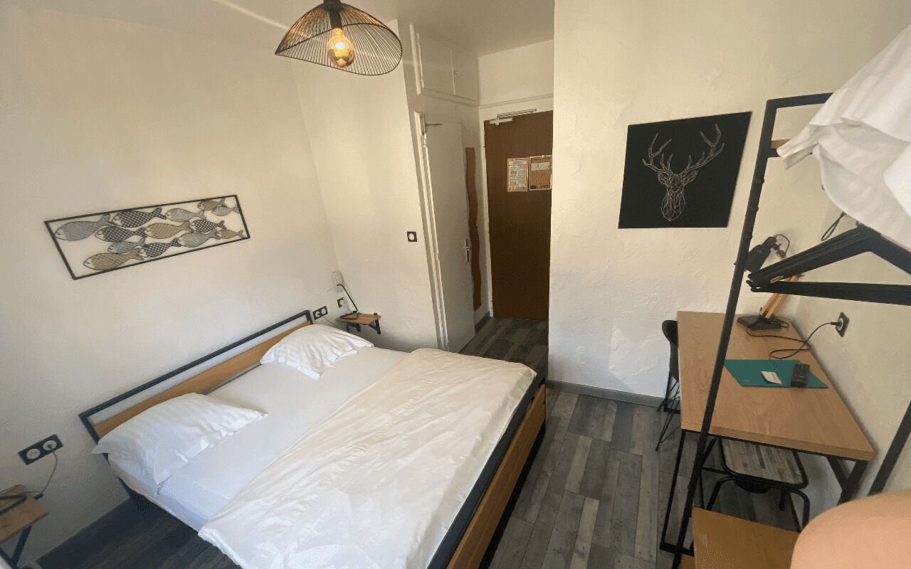 Un lit bien fait, avec du linge de lit blanc, un bureau avec une lampe et une décoration murale représentant une tête de cerf.