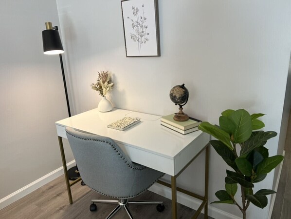 A white desk with a globe and a book, a grey chair, a small plant, and a framed botanical print on the wall.