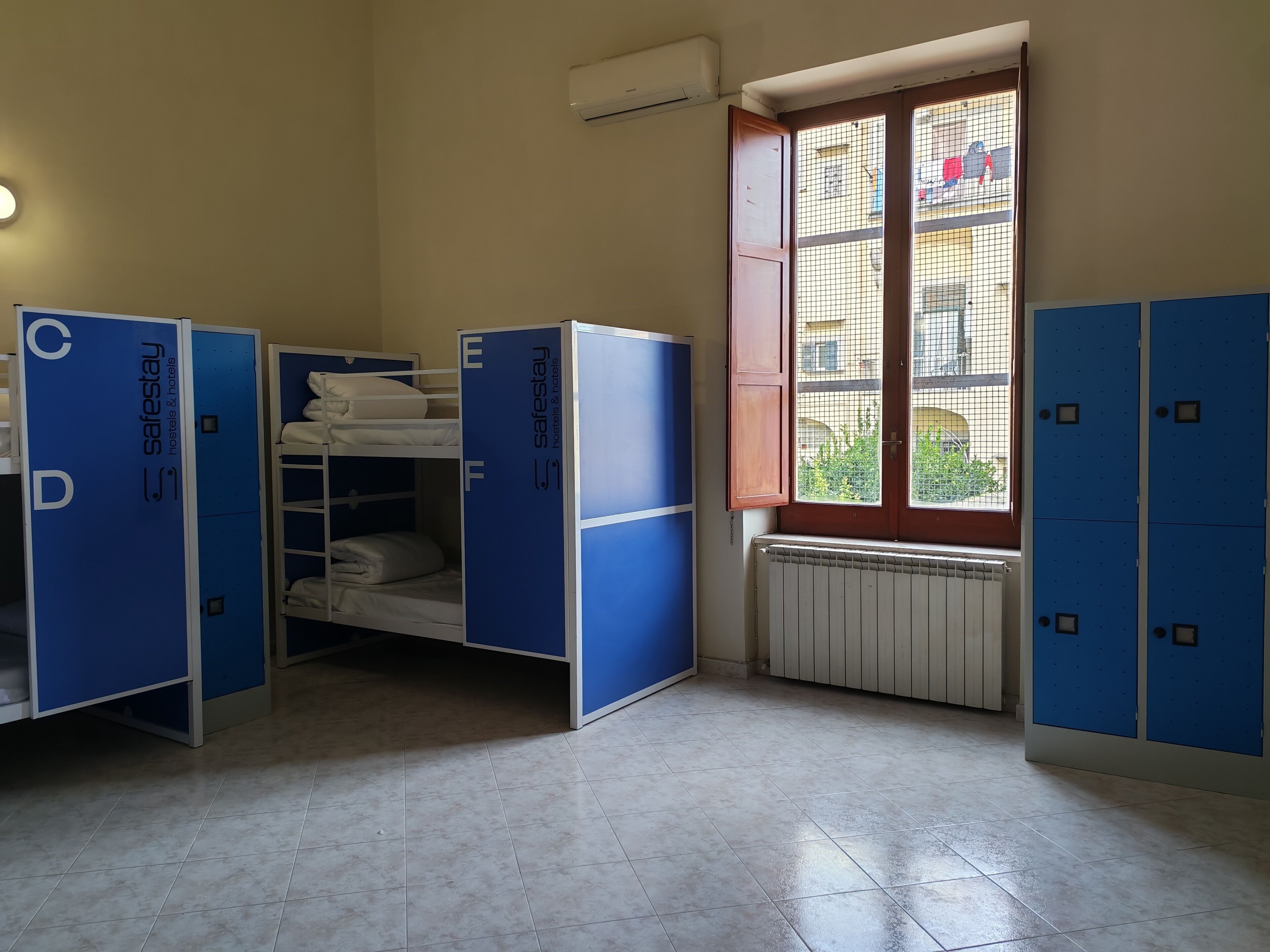 A dormitory room with bunk beds, a radiator, a window with a view of buildings, and blue lockers.