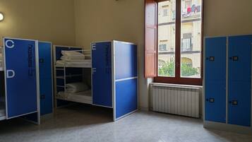 A dormitory room with bunk beds, a radiator, a window with a view of buildings, and blue lockers.