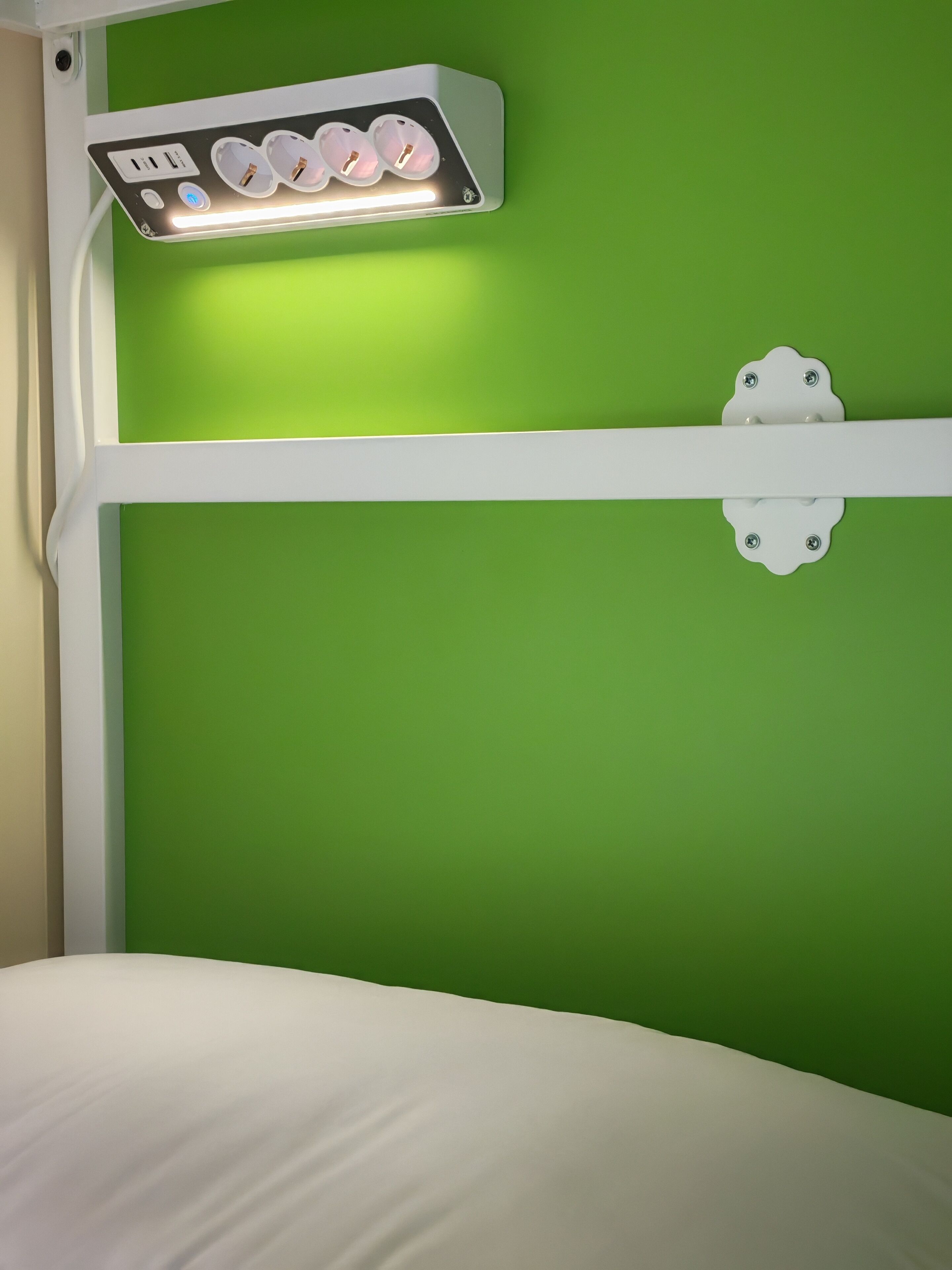 A bed with white bedding, a green wall, and a wall-mounted light fixture with two sockets.