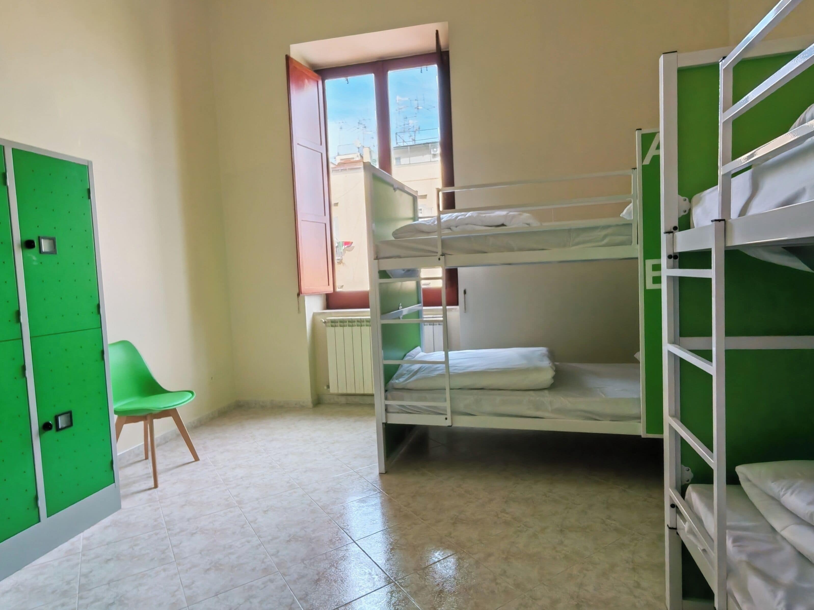 Une chambre de dortoir avec des lits superposés, un radiateur, une chaise verte et une fenêtre donnant sur des immeubles.