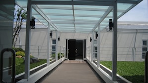 Une allée couverte menant à l’entrée d’un bâtiment, surmontée d’un toit de verre et bordée de verdure et d’une clôture métallique.