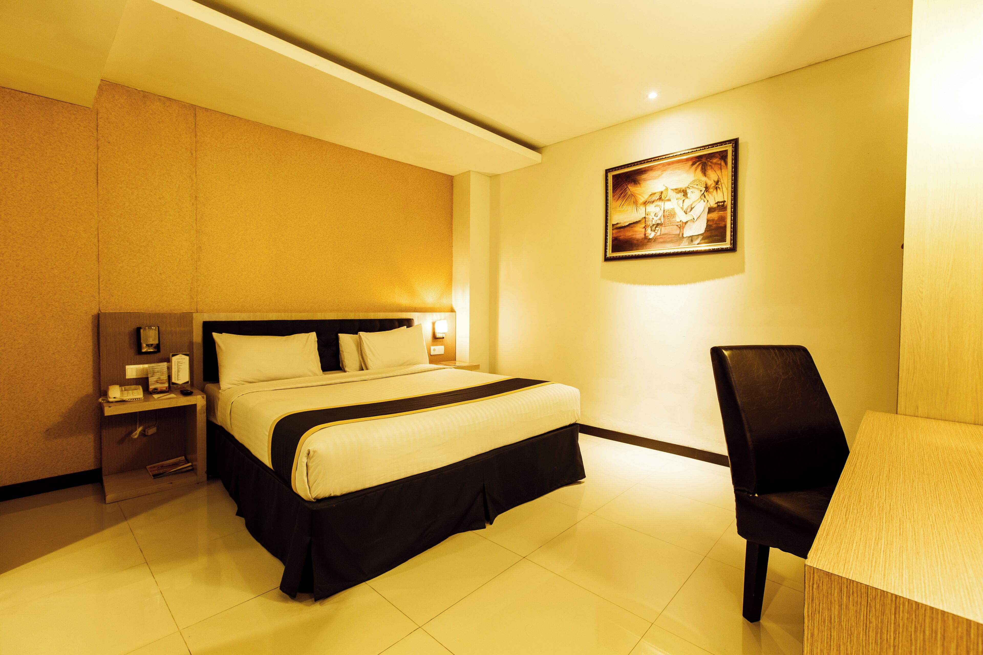A hotel room with a bed, a desk, and a chair. There is a nightstand with a lamp and a picture frame.