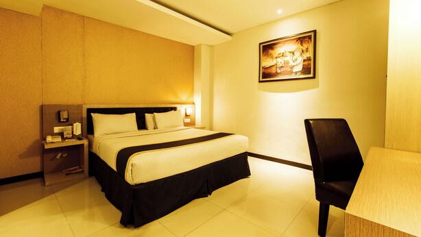 Ein Hotelzimmer mit Bett, Schreibtisch und Stuhl. Ein Nachttisch mit Lampe und Bilderrahmen sind vorhanden.