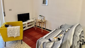 Ein kompaktes Hotelzimmer mit gelbem Sessel, einem Bett mit gestreifter Bettwäsche, einem Fernseher, einem kleinen Holztisch mit Fernbedienung und zwei gerahmten Bildern an der Wand.