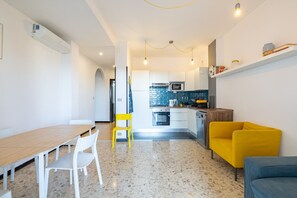Cocina moderna con armarios blancos, microondas y estufa. Hay una zona de comedor con mesa de madera y sillas blancas.