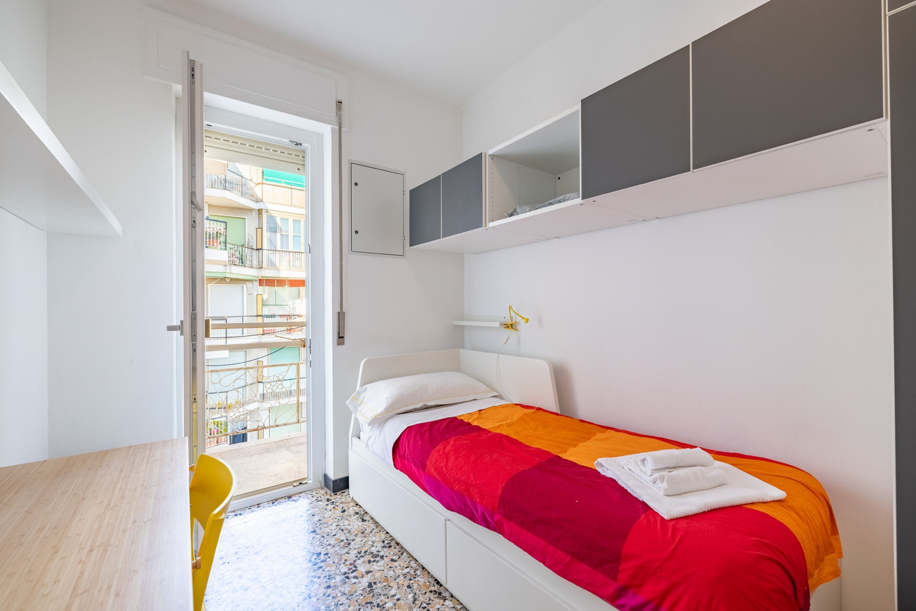 Quarto com cama, escrivaninha e cadeira. Tem uma varanda com guarda-corpo e vista para edifícios.