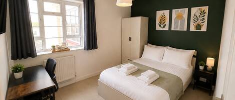 Un dormitorio con cama, escritorio, silla, radiador, ventana con cortinas, armario y decoración en la pared.