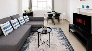Een moderne woonkamer met een grijze bank, een salontafel, een televisie en een eethoek met stoelen en een tafel.