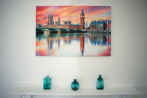 A painting of a cityscape with a bridge and buildings reflected in a body of water, displayed above a white shelf with three glass vases.