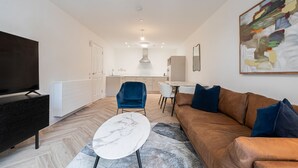 Una sala de estar moderna con un sofá, una televisión, una mesa de centro de mármol y una cocina al fondo.