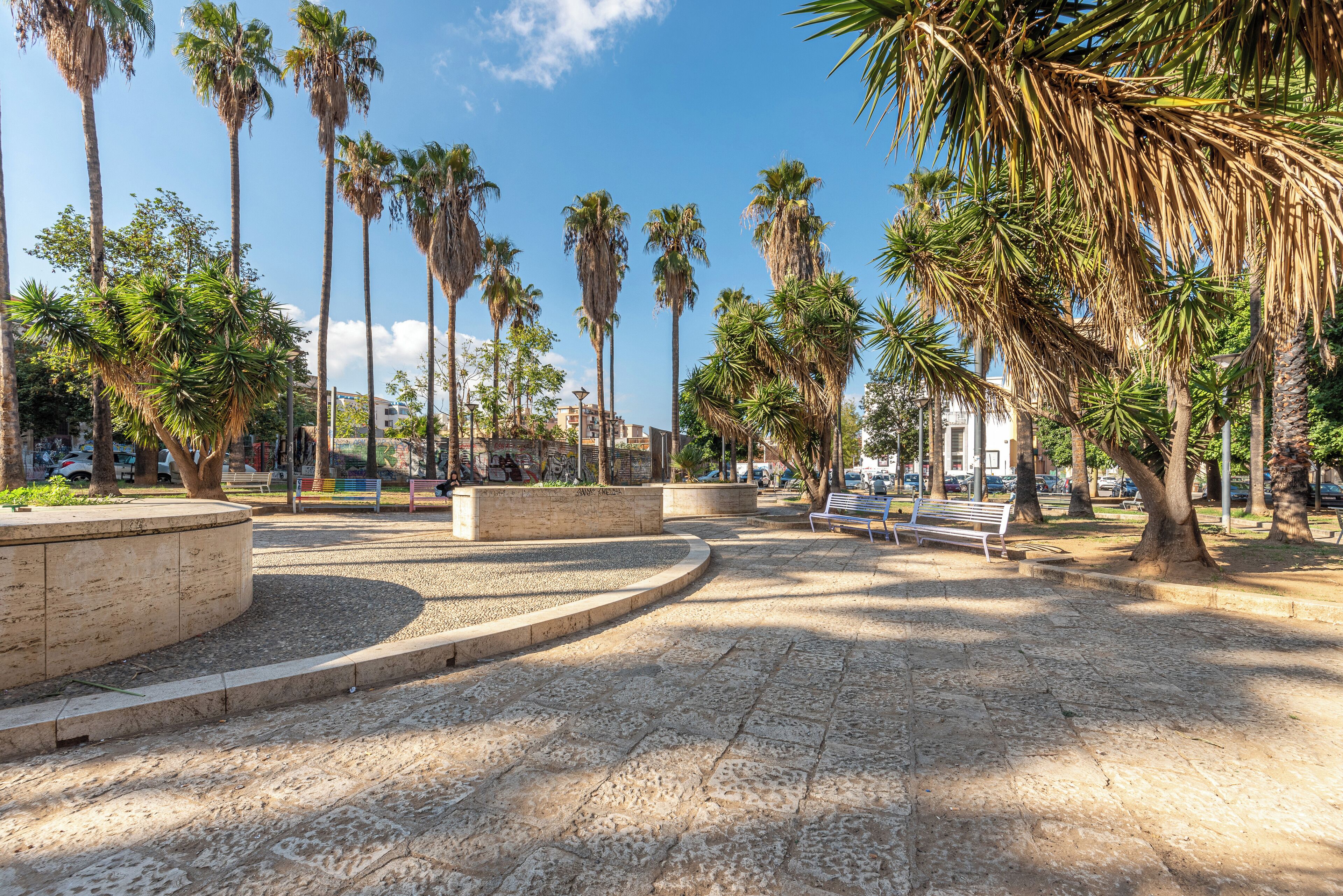 Een verhard park met palmbomen, banken en een ronde stenen constructie.
