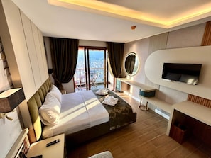 A modern hotel room with a large bed, a TV, a balcony with a city view, and a desk with a mirror. - Castello By Mediterra  (Alanya)