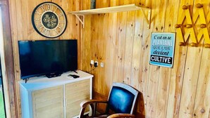 Una habitación con pared de madera, un televisor sobre un mueble blanco, una silla y un letrero que dice “C'est en se PLANTANT QUE L'ON devient CULTIVÉ”.