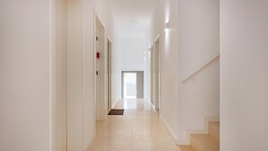 A long, narrow hallway with white walls and a tiled floor, leading to a door at the end.