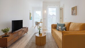A modern living room with a flat-screen TV on a wooden console, a round coffee table with a vase, a sofa with patterned cushions, and a view of a balcony through a large window.