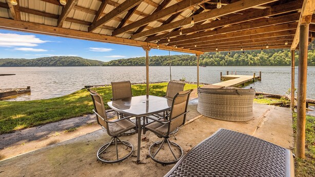 A covered patio with a table and chairs, overlooking a lake with a dock and greenery in the background. - Lakefront retreat with private dock, open living, fire pit, full kitchen & cozy indoor spaces (Radford)