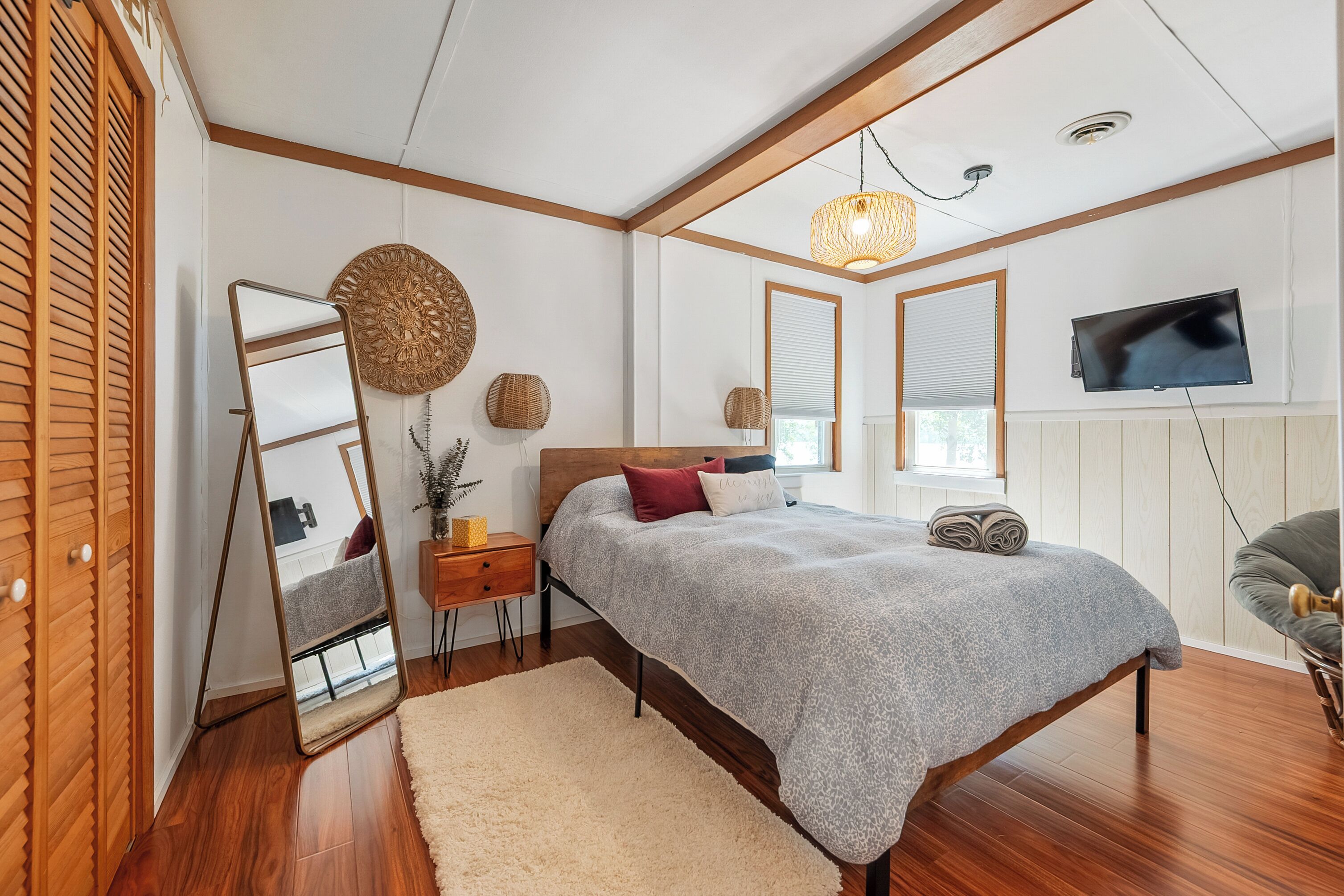 A bedroom with a bed, bedside tables, a mirror, a TV, and a window with blinds.