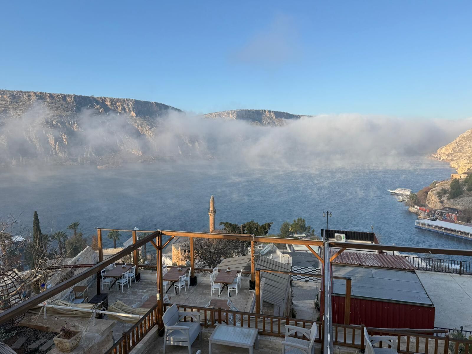 A terrace with outdoor seating overlooking a body of water, a distant mountain, and a boat.