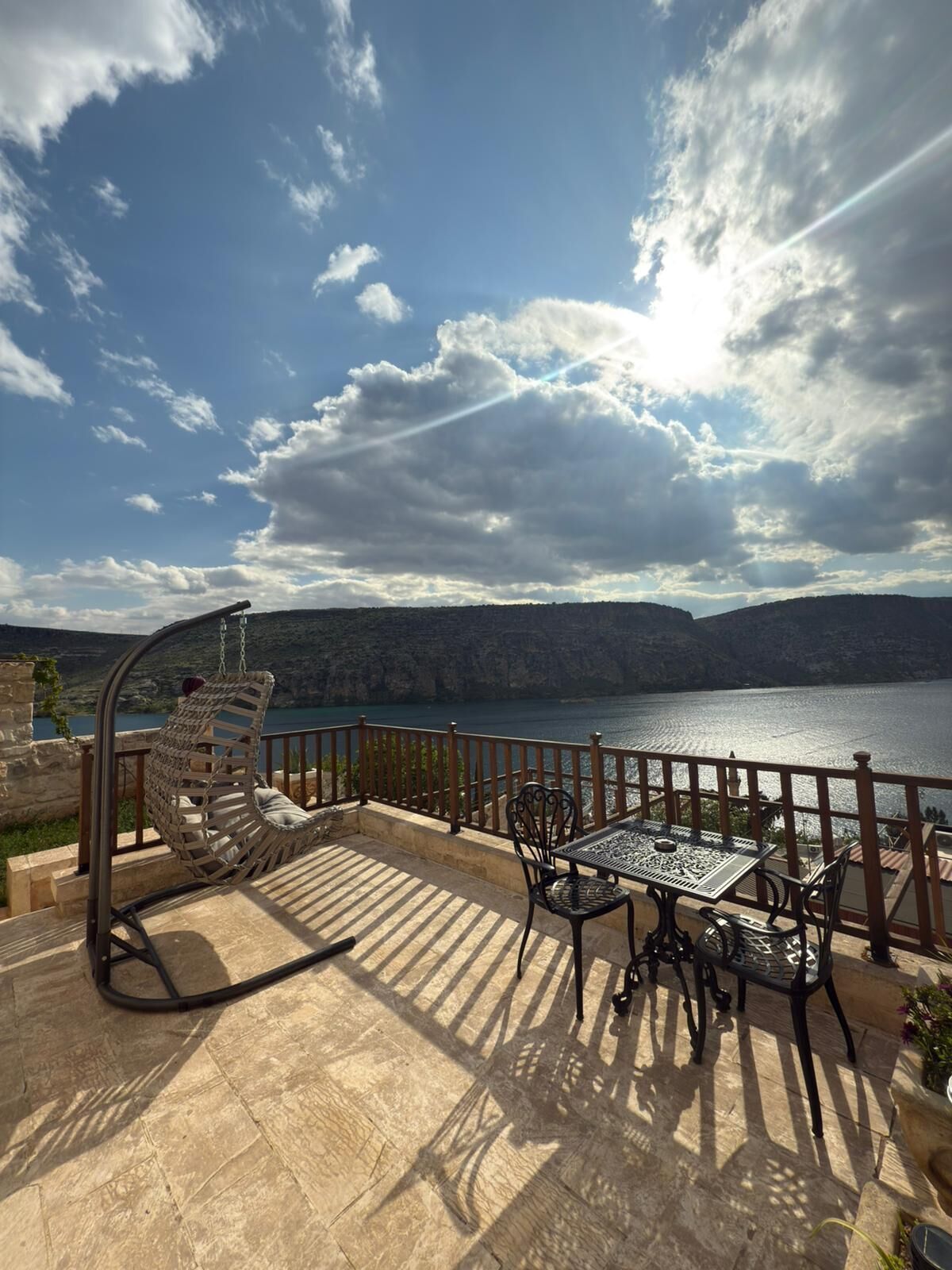 Uma varanda com uma cadeira suspensa, uma mesa e cadeiras, com vista para um lago e montanhas ao fundo.