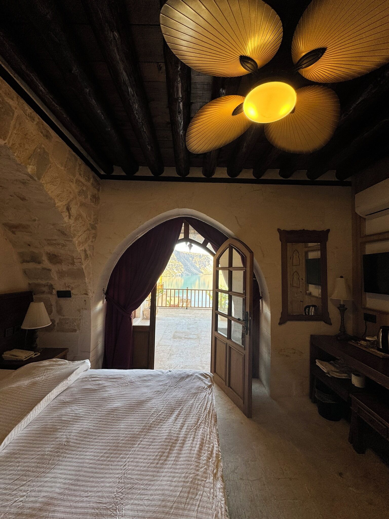 Una habitación con cama, escritorio, espejo y vista a un lago a través de una puerta abierta.