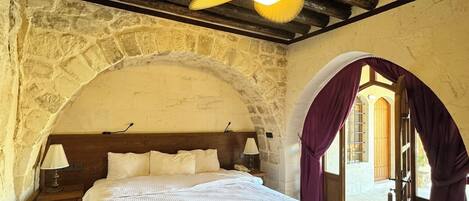 A bedroom with a stone wall, wooden ceiling beams, a bed with white bedding, a nightstand, a lamp, and a ceiling fan.