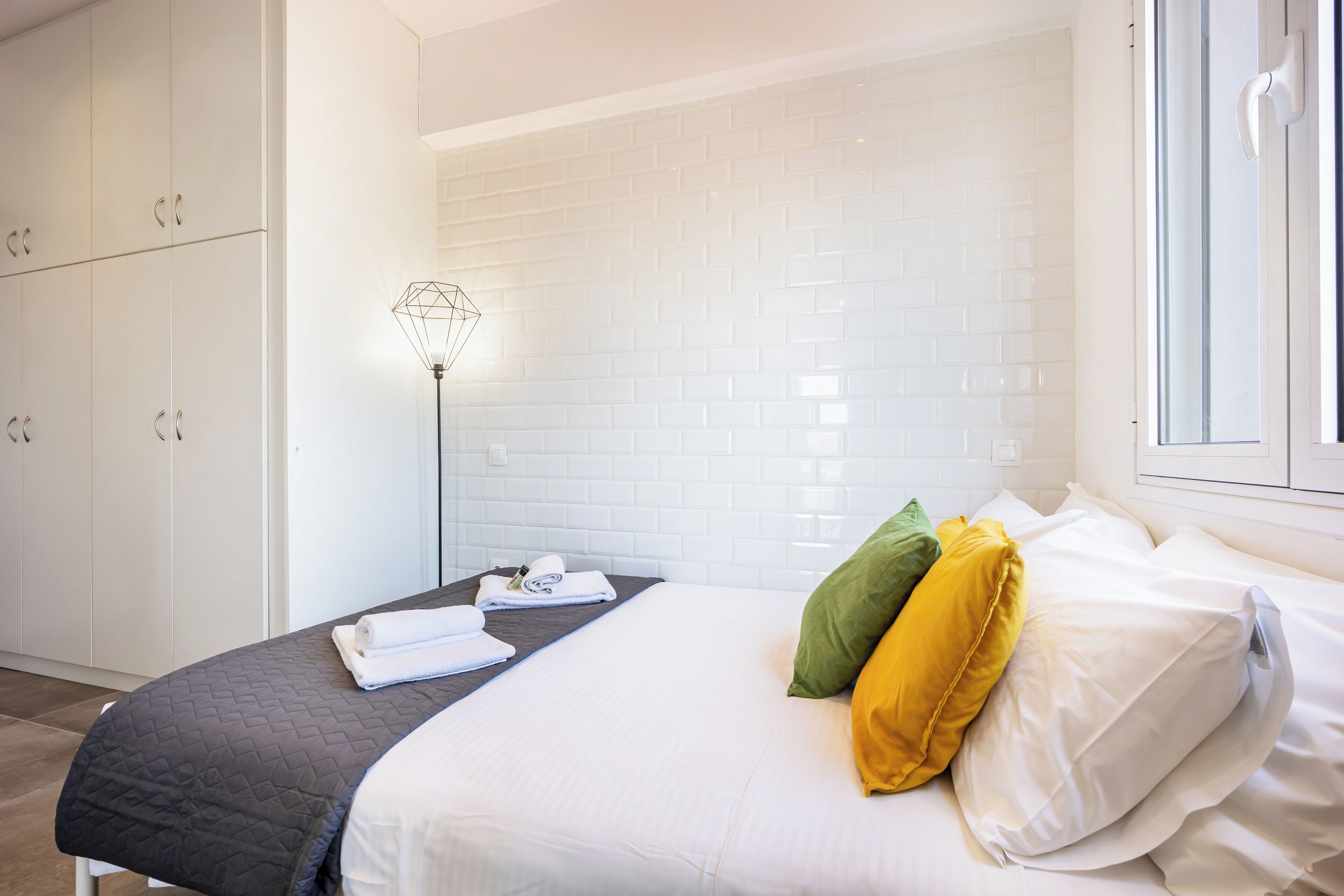 Une chambre d’hôtel avec un lit, des rideaux blancs, un mur recouvert de carreaux blancs et une fenêtre.