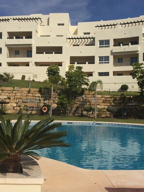 Una piscina con un muro de piedra y palmeras, rodeada por un edificio de varios pisos con balcones.