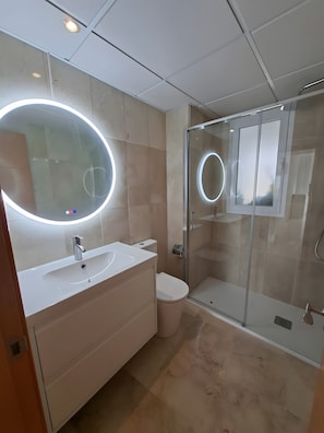 A modern bathroom with a glass shower enclosure, a round mirror with lights, a white sink, and a toilet.