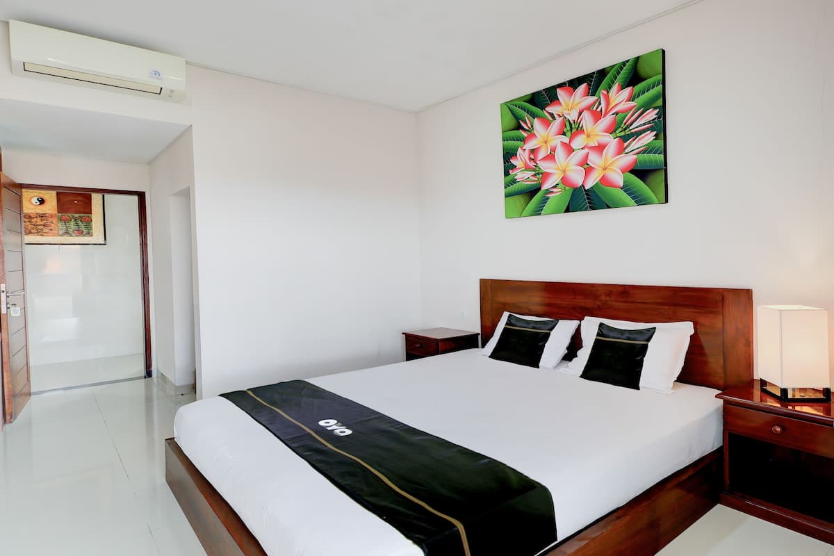 A hotel room with a bed, a nightstand, a wall-mounted air conditioner, and a painting of flowers.