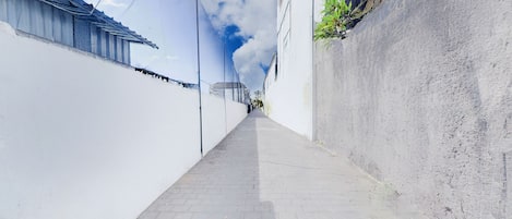 A narrow alleyway with a paved path, flanked by white walls and a blue mesh fence on one side.