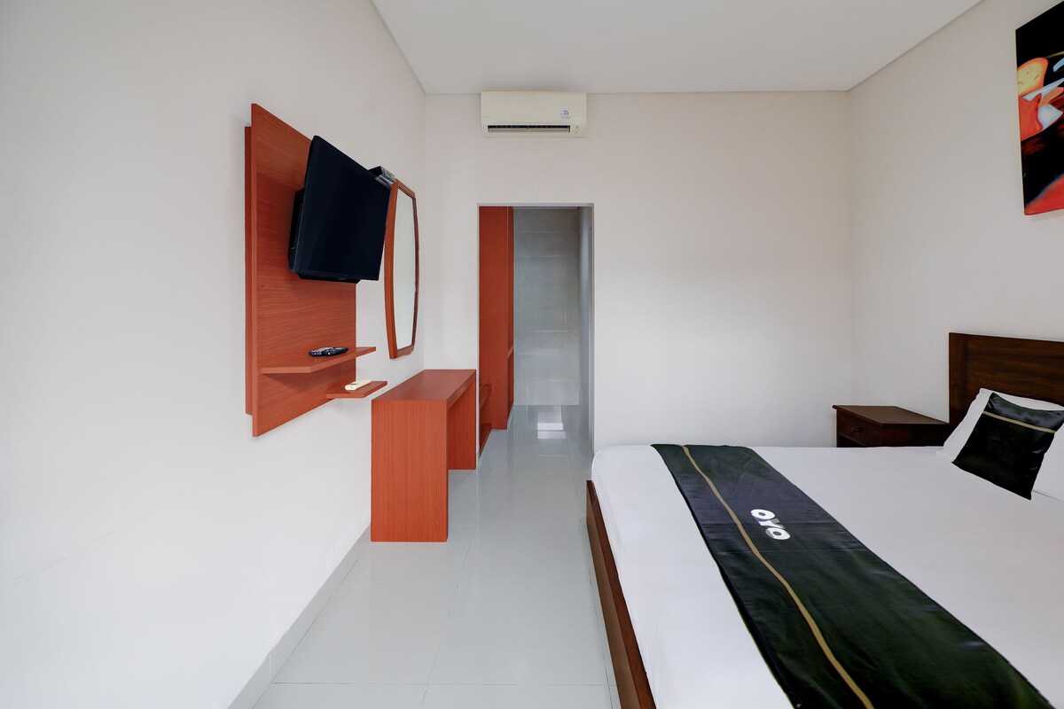 Quarto de hotel com cama, escrivaninha, cadeira, espelho e porta para outro cômodo.