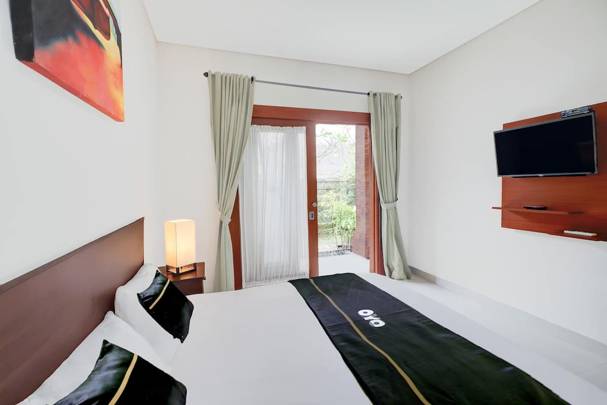 A hotel room with a bed, a TV mounted on the wall, a bedside table with a lamp, and a view of the outdoors through a door.
