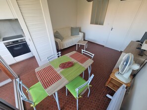 A compact kitchen with a built-in oven, a dining area with a table and chairs, a small bedside table, and a fan.