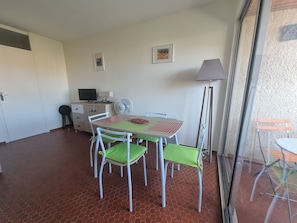 Een kamer met een eettafel, stoelen, een lamp, een televisie en een ventilator.