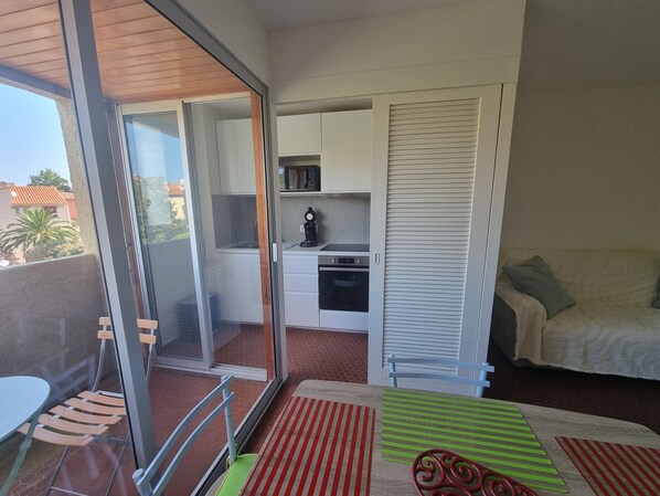 A compact living space with a sliding glass door leading to a balcony, a kitchenette with a microwave and oven, a dining area with a table and chairs, and a bed with a green-striped pillow.