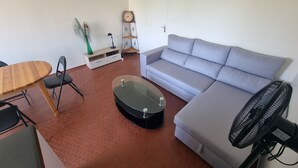 A living room with a grey sofa, a wooden dining table with chairs, a glass coffee table, a black fan, a decorative clock, and a small plant. - Collioure: T2 Apartment with Terrace and Parking, Sleeps 4 (Collioure)