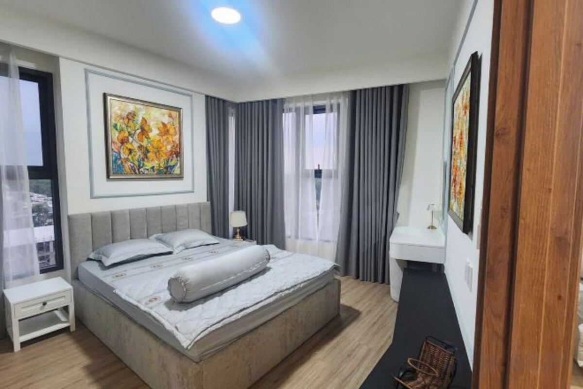 A hotel room with a large bed, a bedside table, a nightstand, a window with curtains, and a painting on the wall.