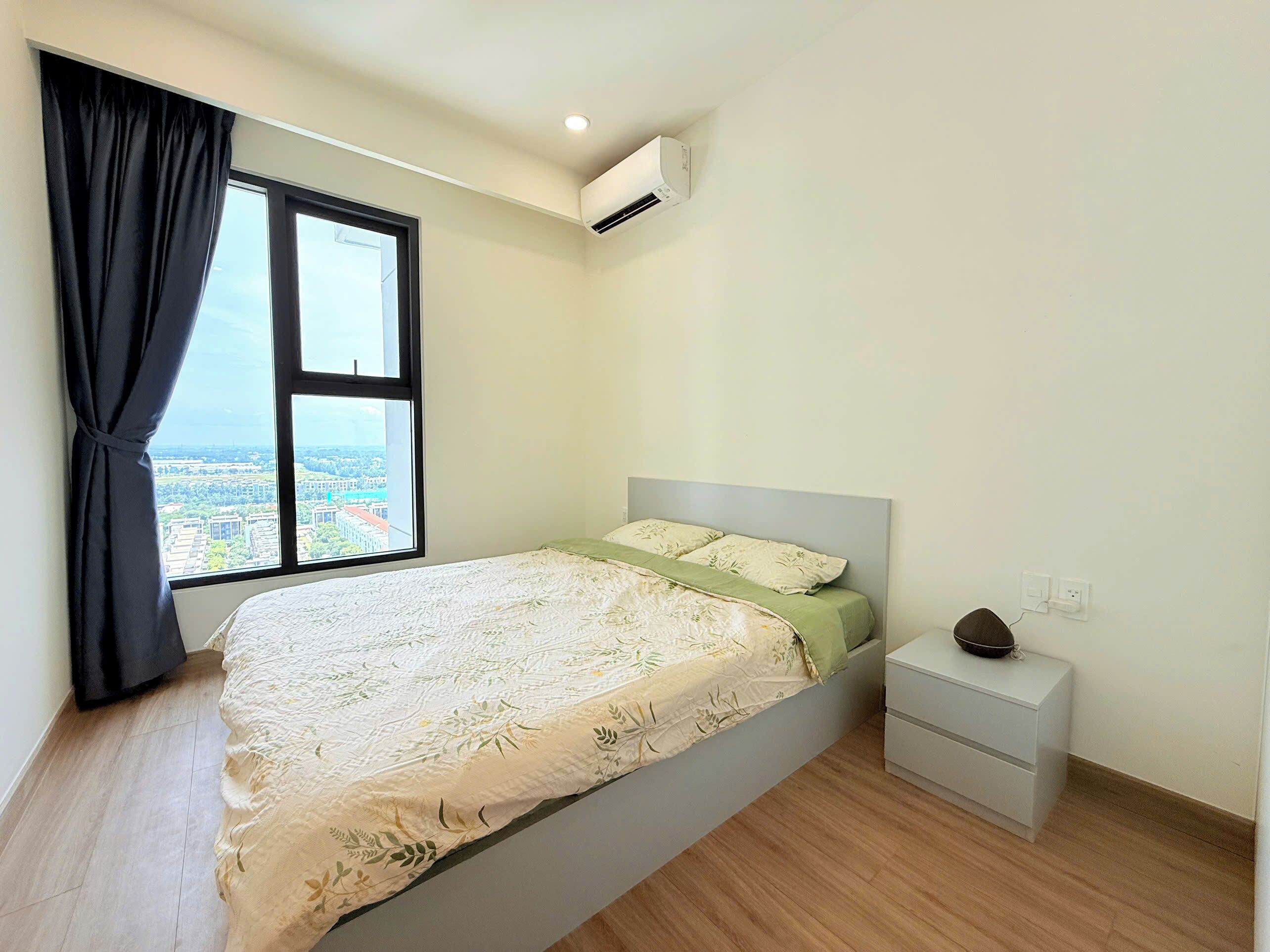 A bedroom with a bed, a nightstand, a window with a view of buildings, and an air conditioning unit.