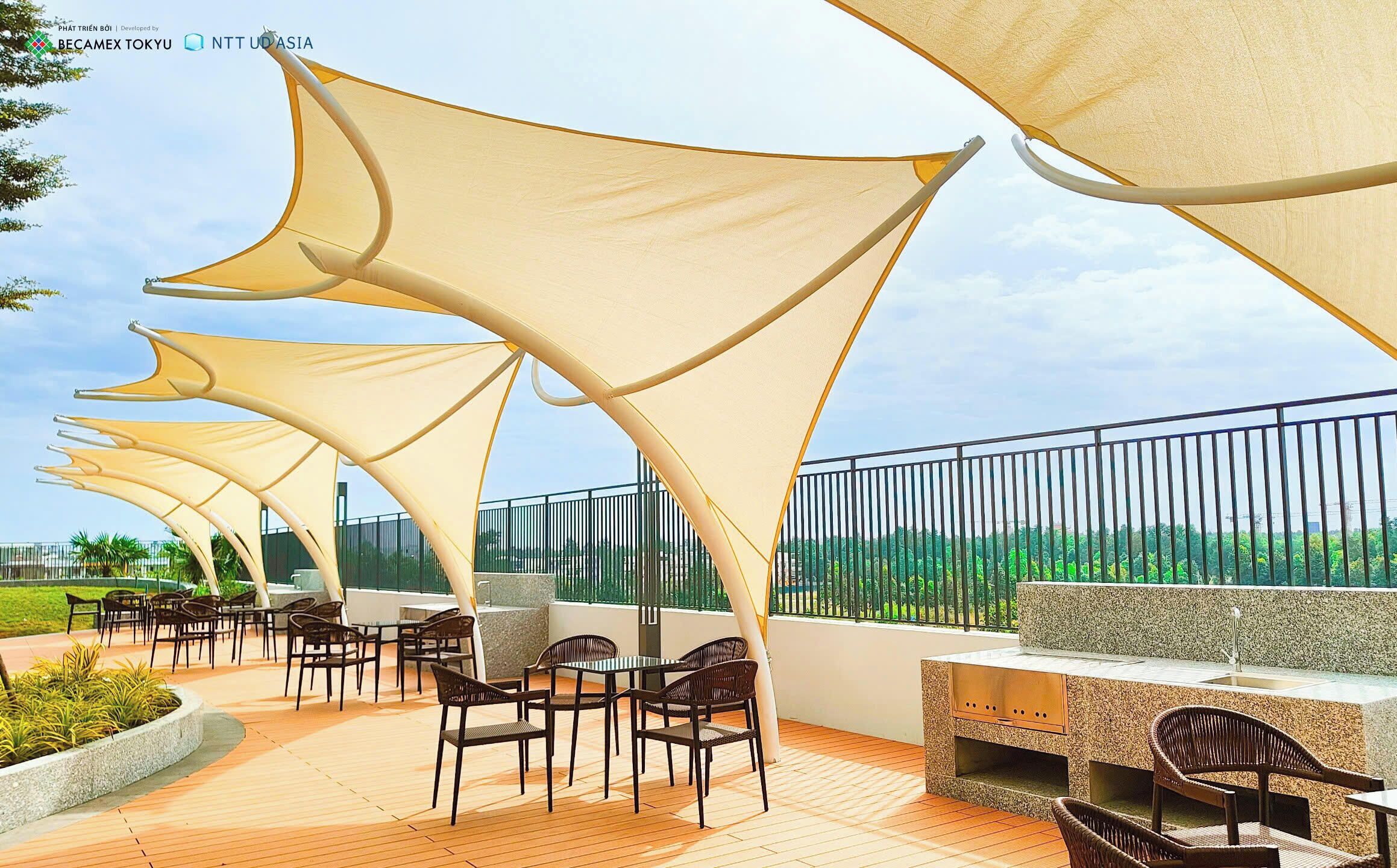 An outdoor seating area with tables and chairs, shaded by large fabric canopies, and a view of greenery in the background.