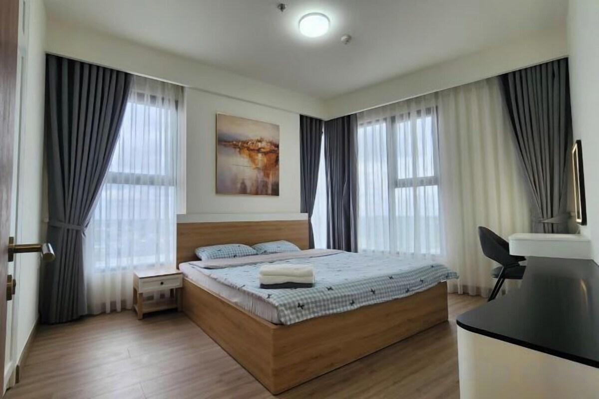 A bedroom with a large bed, a desk, and a chair. There is a window with curtains and a painting on the wall.